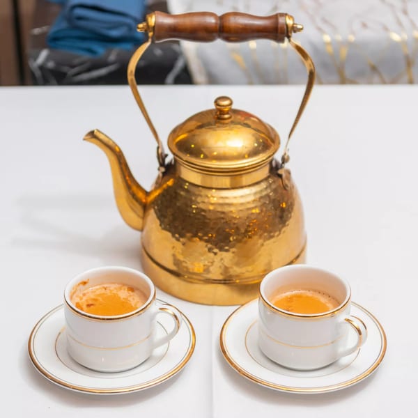 A golden teapot and two white cups of Indian chai tea on a white tablecloth.
