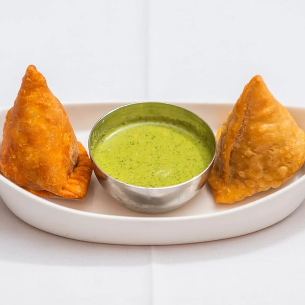 Two golden-brown samosas flank a silver bowl of vibrant green chutney on a white plate.