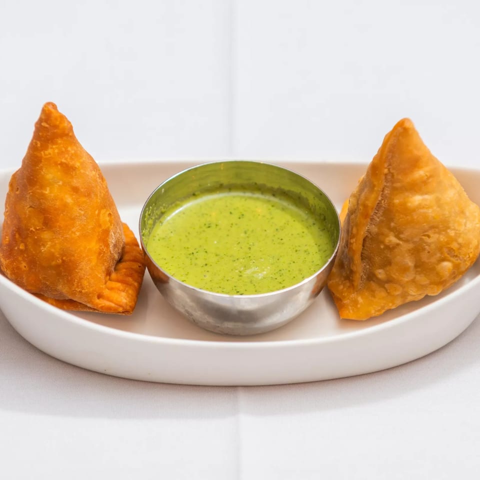 Two golden-brown samosas flank a silver bowl of vibrant green chutney on a white plate.
