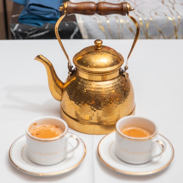 A golden kettle and two small white cups of chai tea on a white tablecloth.