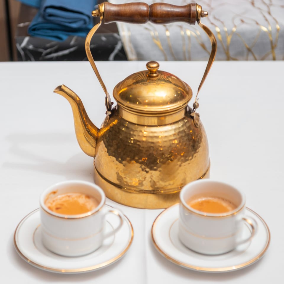 A golden kettle and two small white cups of chai tea on a white tablecloth.