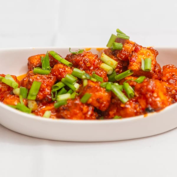 Close-up of vibrant red chili paneer, garnished with fresh green scallions in a white bowl.
