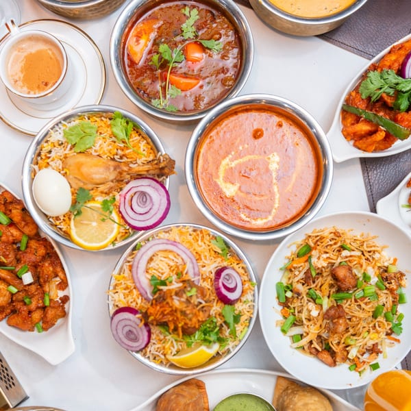 Overhead view of various Indian dishes including biryani, curries, noodles, and chai on a white table.