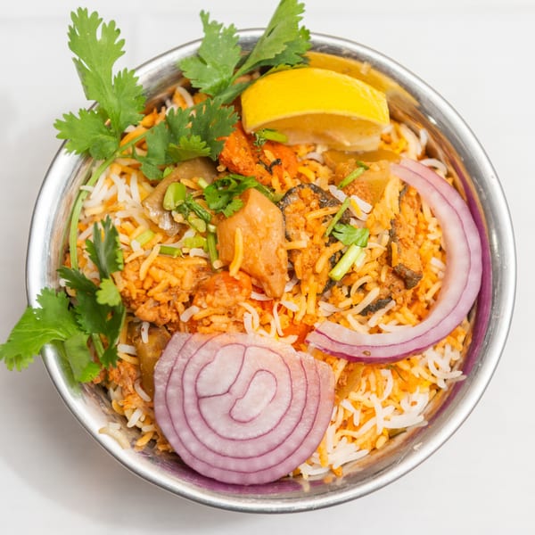 A metal bowl of King of Tandoor & Biryani House's biryani, garnished with cilantro, lemon, and red onion.