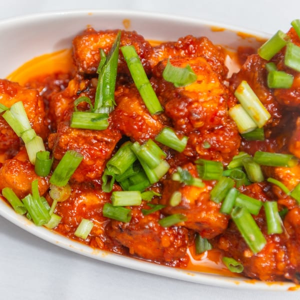 Close-up of vibrant orange Indian chili paneer, garnished with fresh green scallions in a white bowl.