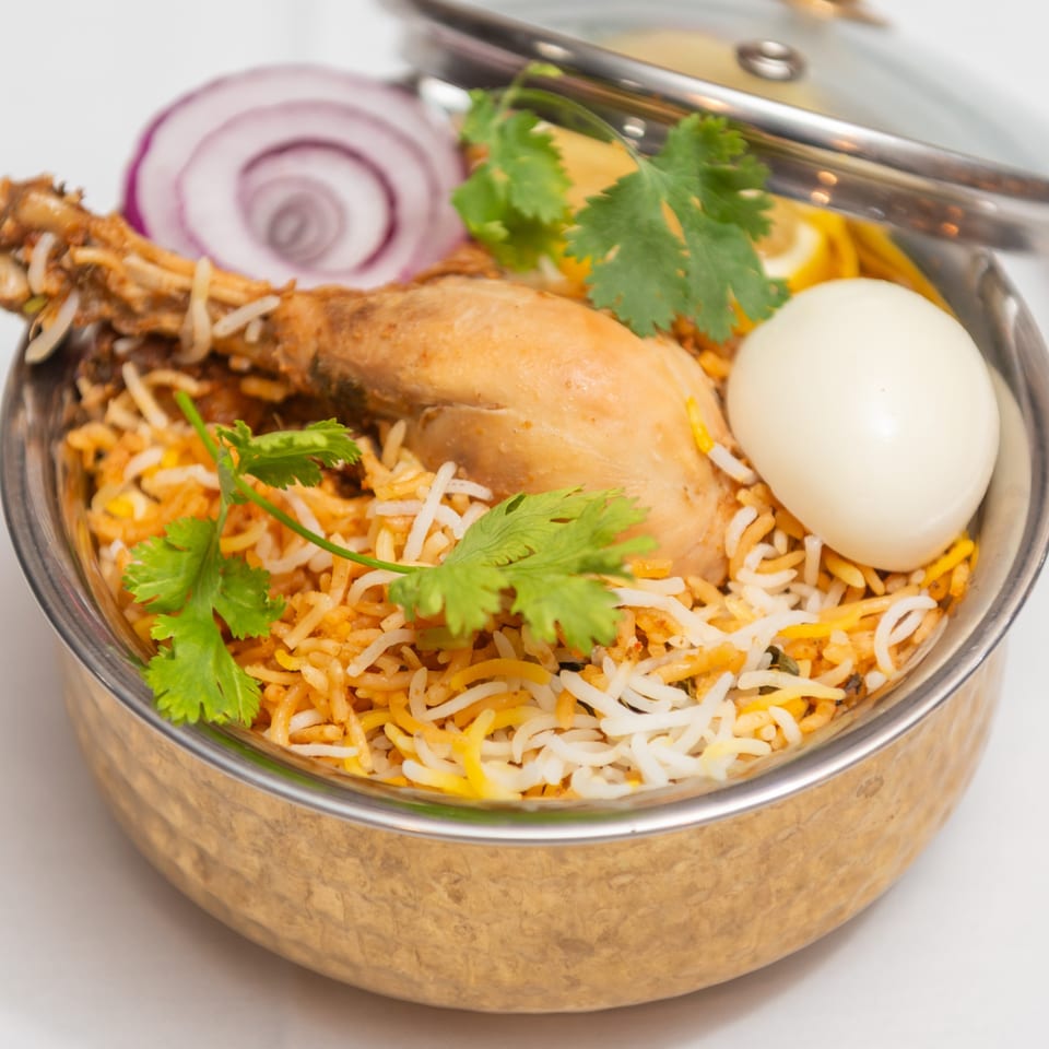 Chicken biryani with a boiled egg, red onion, and cilantro in a brass bowl with a lid.