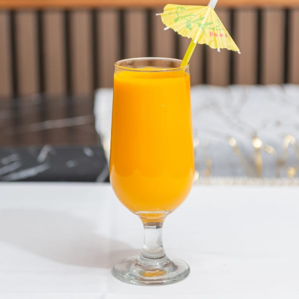 A tall glass of vibrant orange mango lassi with a yellow paper umbrella and straw.