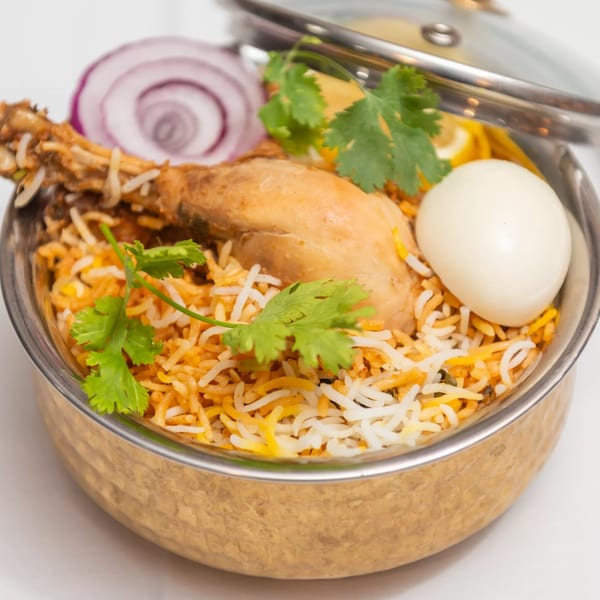 Chicken biryani in a metal bowl, garnished with cilantro, red onion, lemon, and a hard-boiled egg.
