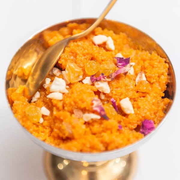 Close-up of orange Indian halwa dessert in a gold bowl, garnished with nuts, raisins, and rose petals.